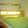 Ein gemütliches Zimmer mit einem Doppelbett, grüner Bettwäsche und einer Wanddekoration mit gelben Blüten und Früchten.