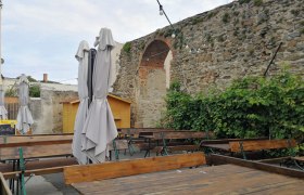 Ein leerer Außenbereich eines Heurigen mit Holztischen, geschlossenen Sonnenschirmen und einer alten Steinmauer im Hintergrund.