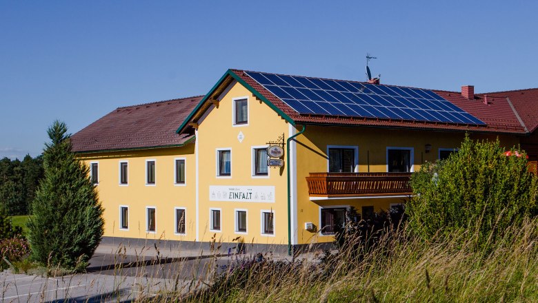 Gelbes Gebäude mit Solarpanelen auf dem Dach, umgeben von grüner Landschaft.