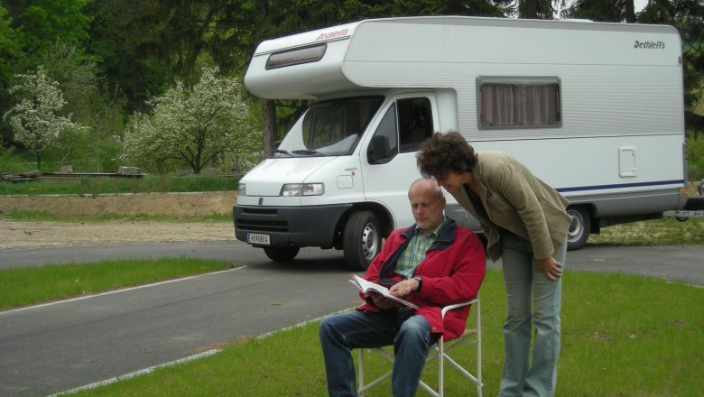 A man sits on a camping chair in front of a camper van while a woman looks over his shoulder. Trees and a meadow can be seen in the background.