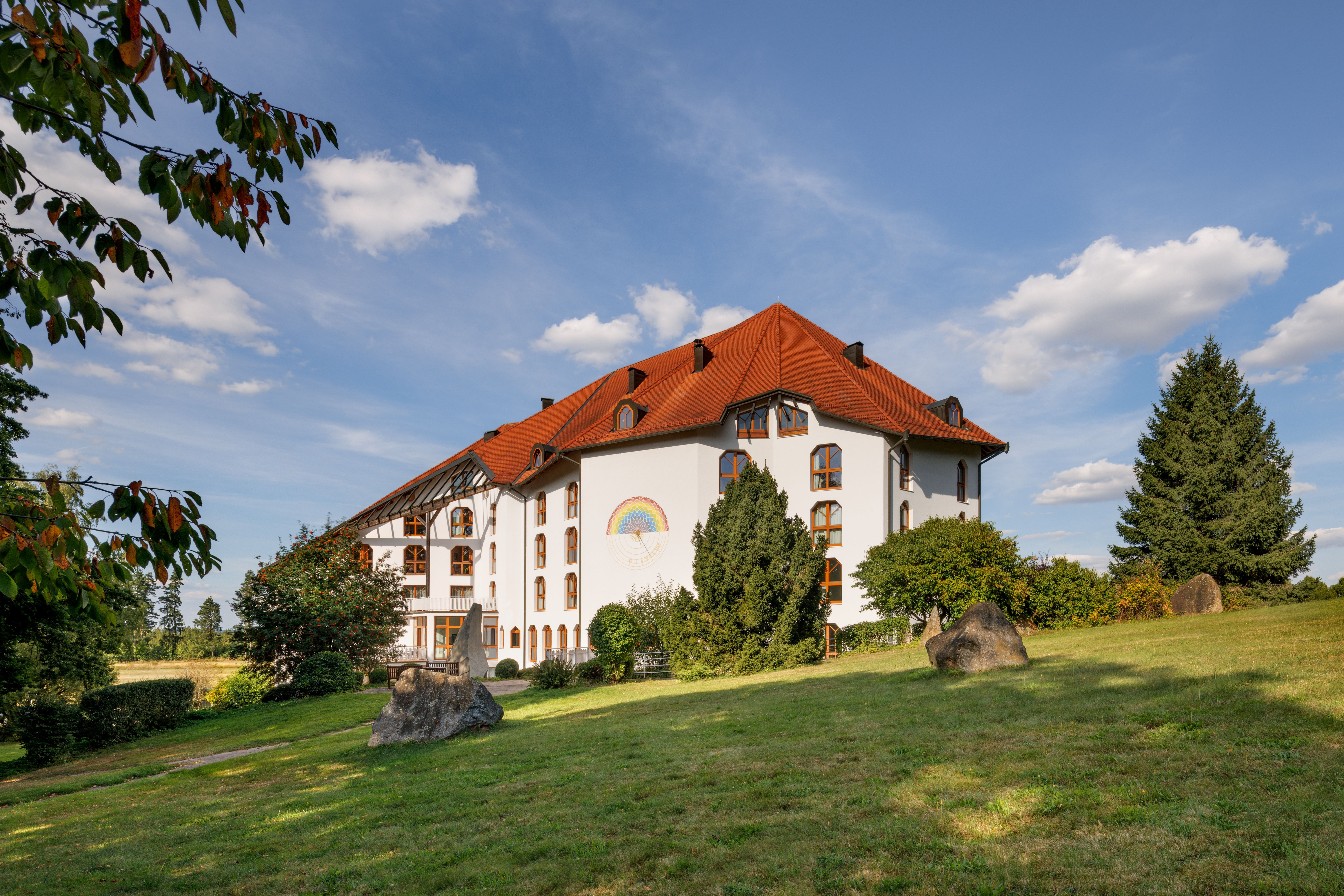 Ein großes, weißes Gebäude mit rotem Dach und einem Regenbogenbild an der Wand, umgeben von grüner Landschaft und Bäumen unter blauem Himmel.