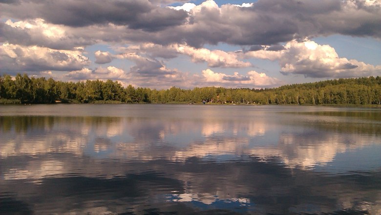 Ein See mit bewölktem Himmel und Wald im Hintergrund.
