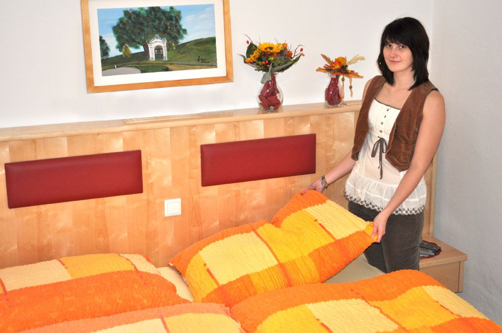 Eine Frau steht in einem Schlafzimmer mit orange-gelber Bettwäsche und Blumenvasen auf dem Kopfteil.