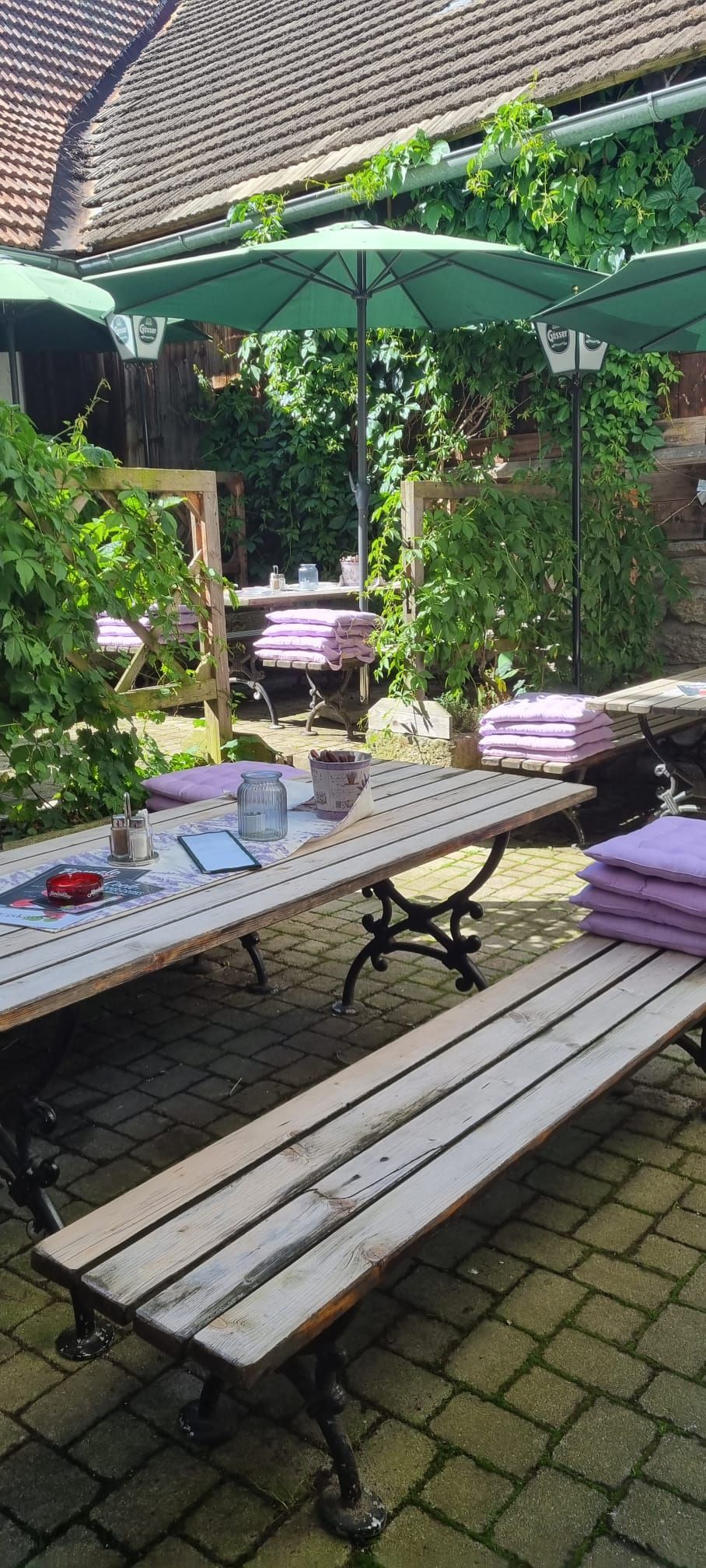 Ein gemütlicher Gastgarten mit Holzbänken, Tischen und grünen Sonnenschirmen, umgeben von Pflanzen und einem Gebäude.