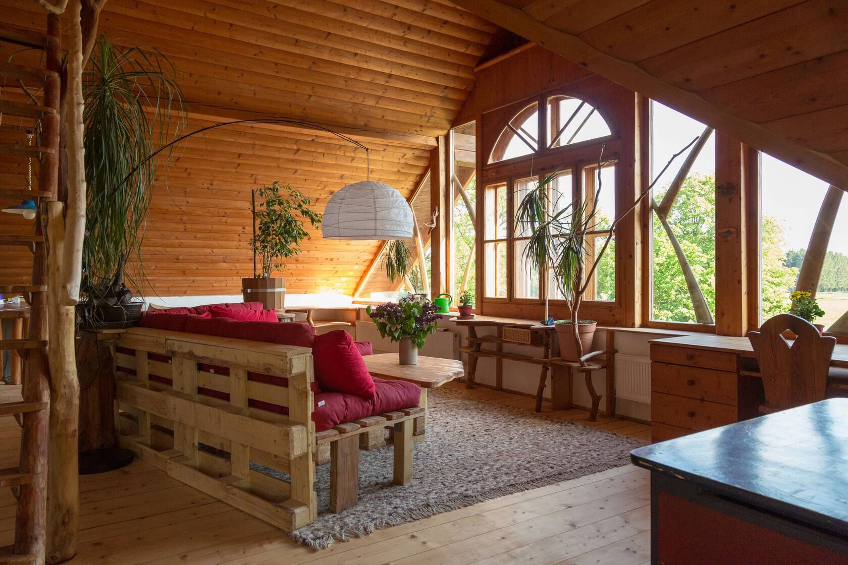 Gemütliches Holzwohnzimmer mit rotem Sofa, großen Fenstern, Pflanzen und Blick ins Grüne.