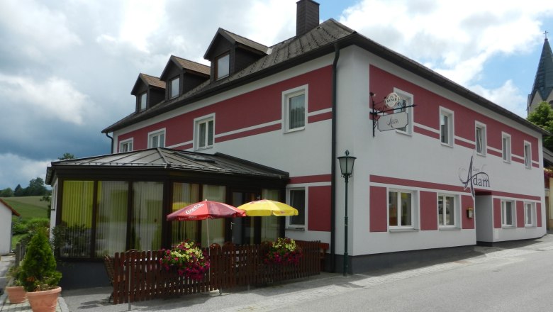 Ein Gasthaus mit roter und weißer Fassade, Terrasse mit Sonnenschirmen und Blumendekoration.