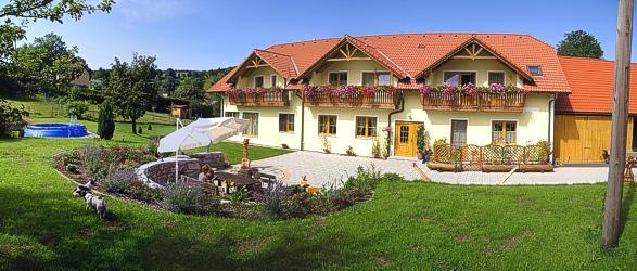 Ein großes gelbes Haus mit roten Dächern und Blumenkästen, umgeben von einem gepflegten Garten mit Sitzbereich und Sonnenschirm.
