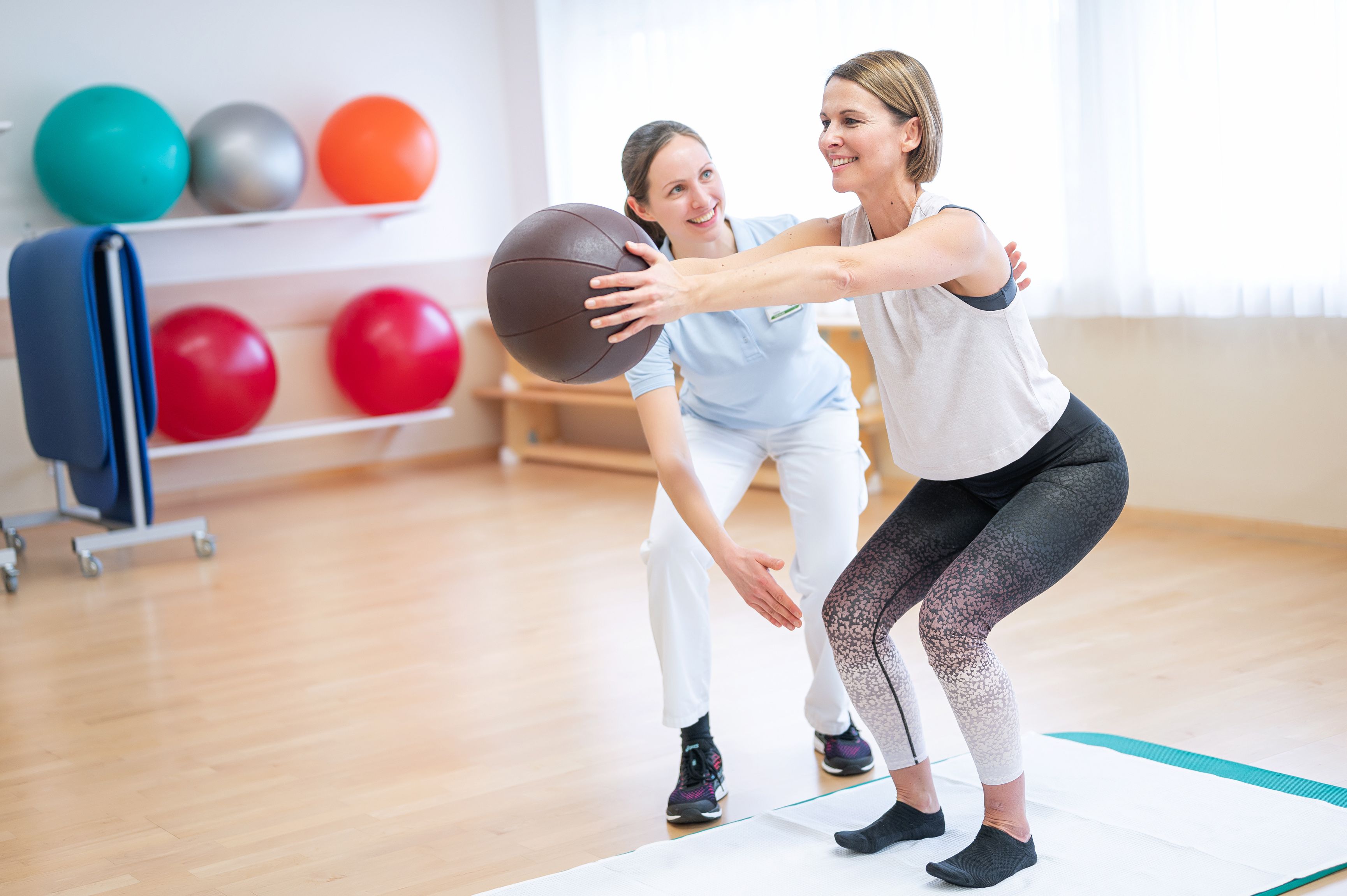Eine Frau macht Gymnastikübungen mit einem Medizinball, während eine Trainerin sie anleitet.