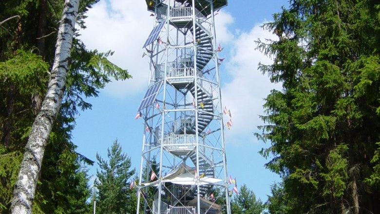 Ein hoher Aussichtsturm aus Metall mit Holzverkleidung, umgeben von Bäumen, unter blauem Himmel.