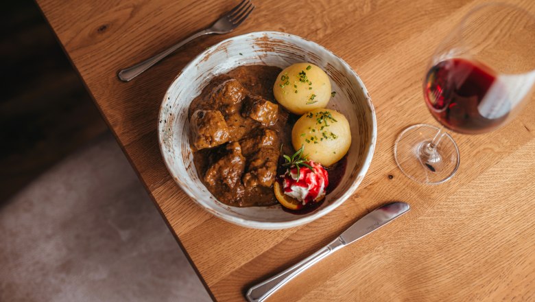A plate of venison goulash, dumplings and cranberries, next to a glass of red wine on a wooden table.