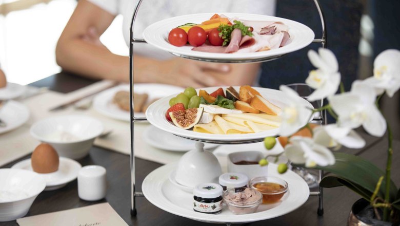 Ein Frühstückstisch mit einer Etagere voller Käse, Aufschnitt, Obst und Brotaufstrichen. Im Vordergrund eine weiße Orchidee.