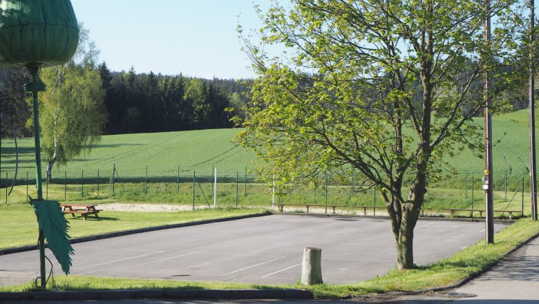 Asphaltplatz mit Baum und Volleyballfeld im Hintergrund.