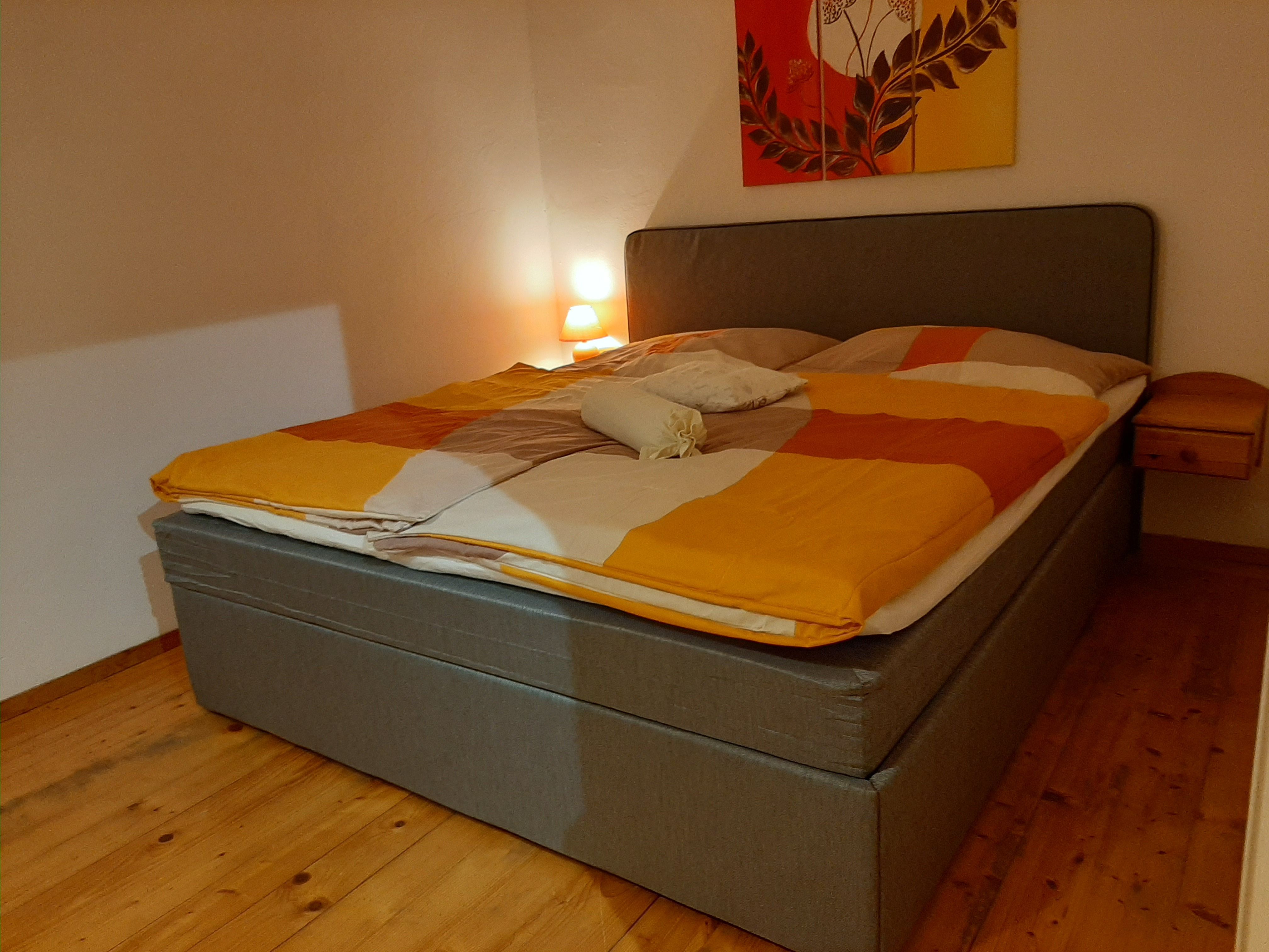 Gemütliches Schlafzimmer mit Doppelbett, grauem Kopfteil, Nachttischlampe und farbig gemusterter Bettdecke.