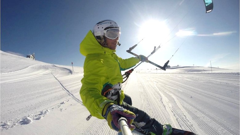 Person beim Snowkiten auf einer verschneiten Piste mit blauem Himmel und Sonnenschein.