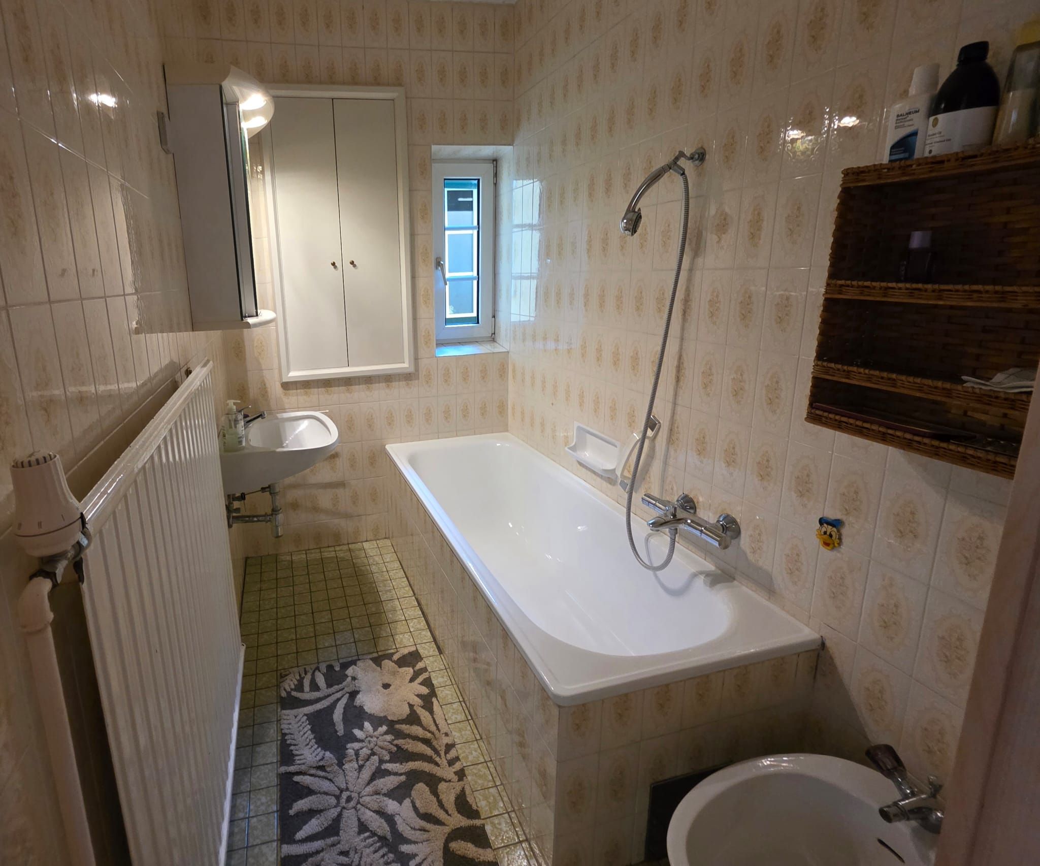 Ein kleines Badezimmer mit Badewanne, Waschbecken, kleinem Fenster und Fliesen an den Wänden. Ein Heizkörper und ein Regal sind ebenfalls sichtbar.
