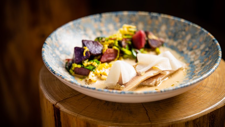 Ein kunstvoll angerichtetes Gericht mit Fischfilet, bunten Kartoffeln und Beurre Blanc in einer dekorativen Schale.