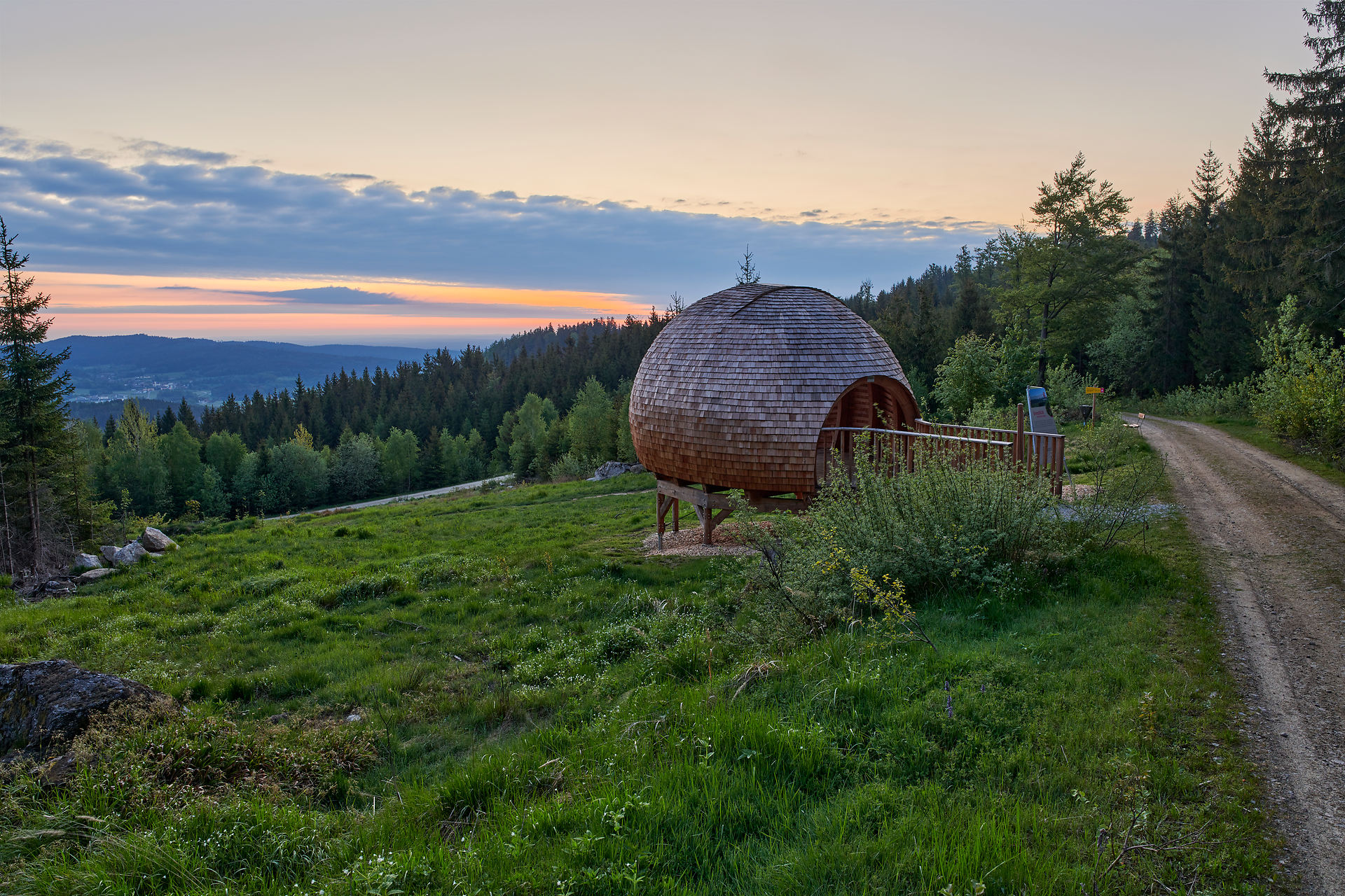 In der sanften Dämmerung erstrahlt die einzigartige Holzkonstruktion in warmen Tönen, umgeben von üppigem Grün und majestätischen Bäumen. Der Weg schlängelt sich durch die malerische Landschaft und lädt Wanderer ein, die atemberaubende Aussicht auf die umliegenden Berge zu genießen.
