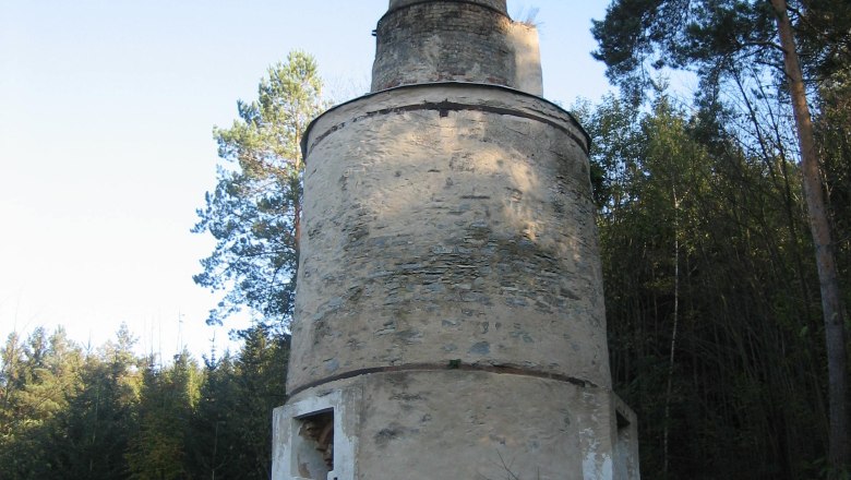 Ein alter, runder Schornstein aus Stein in einem Waldgebiet mit Informationstafel.