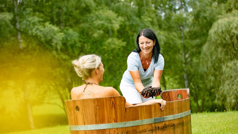 Zwei Frauen bei einer Moorbehandlung im Freien, eine sitzt in einer Holzwanne, die andere reicht ihr Moor.