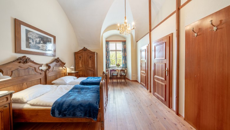 Ein elegantes Doppelzimmer mit antiken Holzm&ouml;beln, einem Kronleuchter und einem Fenster mit Blick ins Gr&uuml;ne.