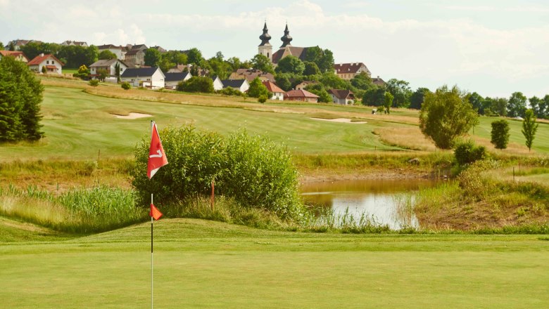Golfplatz mit Fahne im Vordergrund, Dorf und Kirche im Hintergrund.