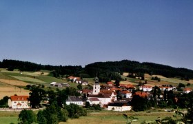 Panorama eines Dorfes mit Kirche und roten Dächern, umgeben von grünen Feldern und Wäldern.
