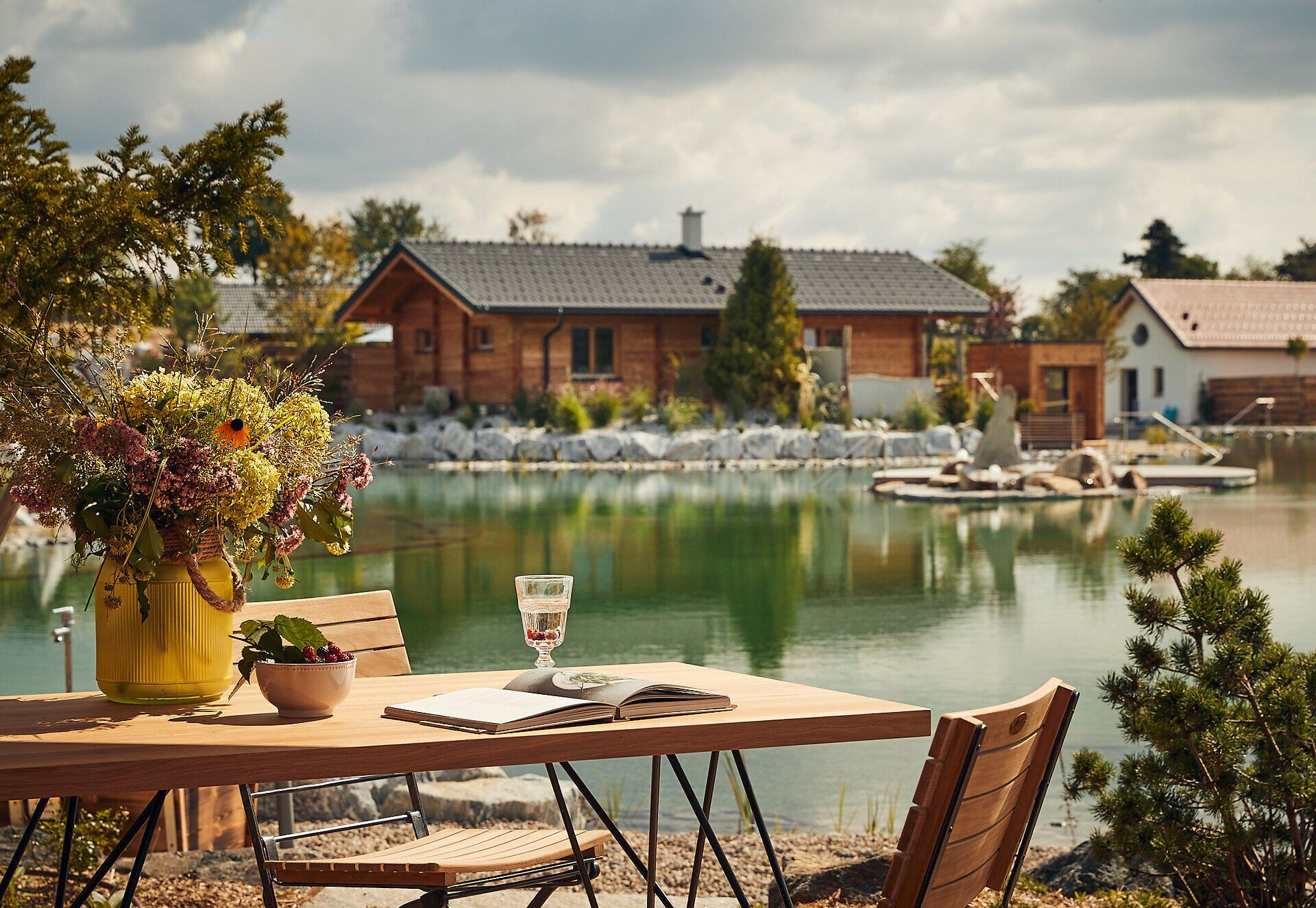 Ein idyllischer Rückzugsort am Gartensee, wo die sanften Wellen des Wassers die umliegenden Bäume spiegeln. Die bunten Blumenarrangements auf dem Tisch laden dazu ein, die Ruhe und Schönheit der Natur zu genießen. Hier wird der Sommer in seiner vollen Pracht erlebbar.