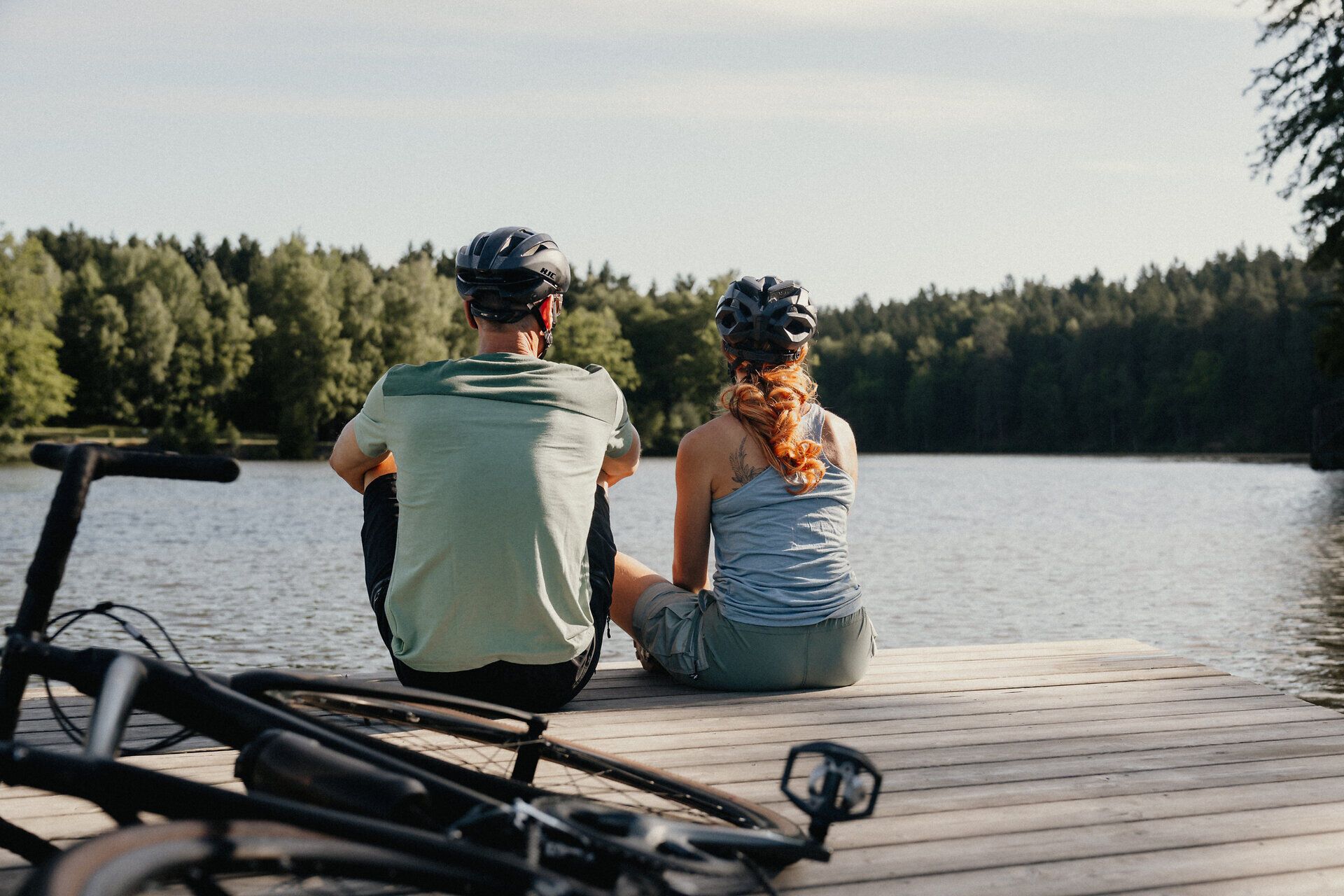 Zwei Radfahrer:innen mit Helmen sitzen an einem Steg und blicken in die Ferne, hinter ihnen die Fahrräder.