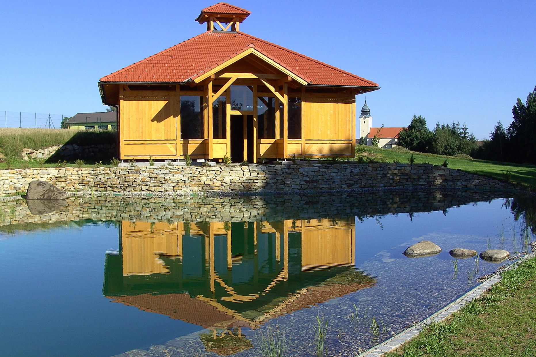 Holzgebäude mit rotem Dach spiegelt sich in einem Teich, umgeben von grüner Landschaft und blauem Himmel.