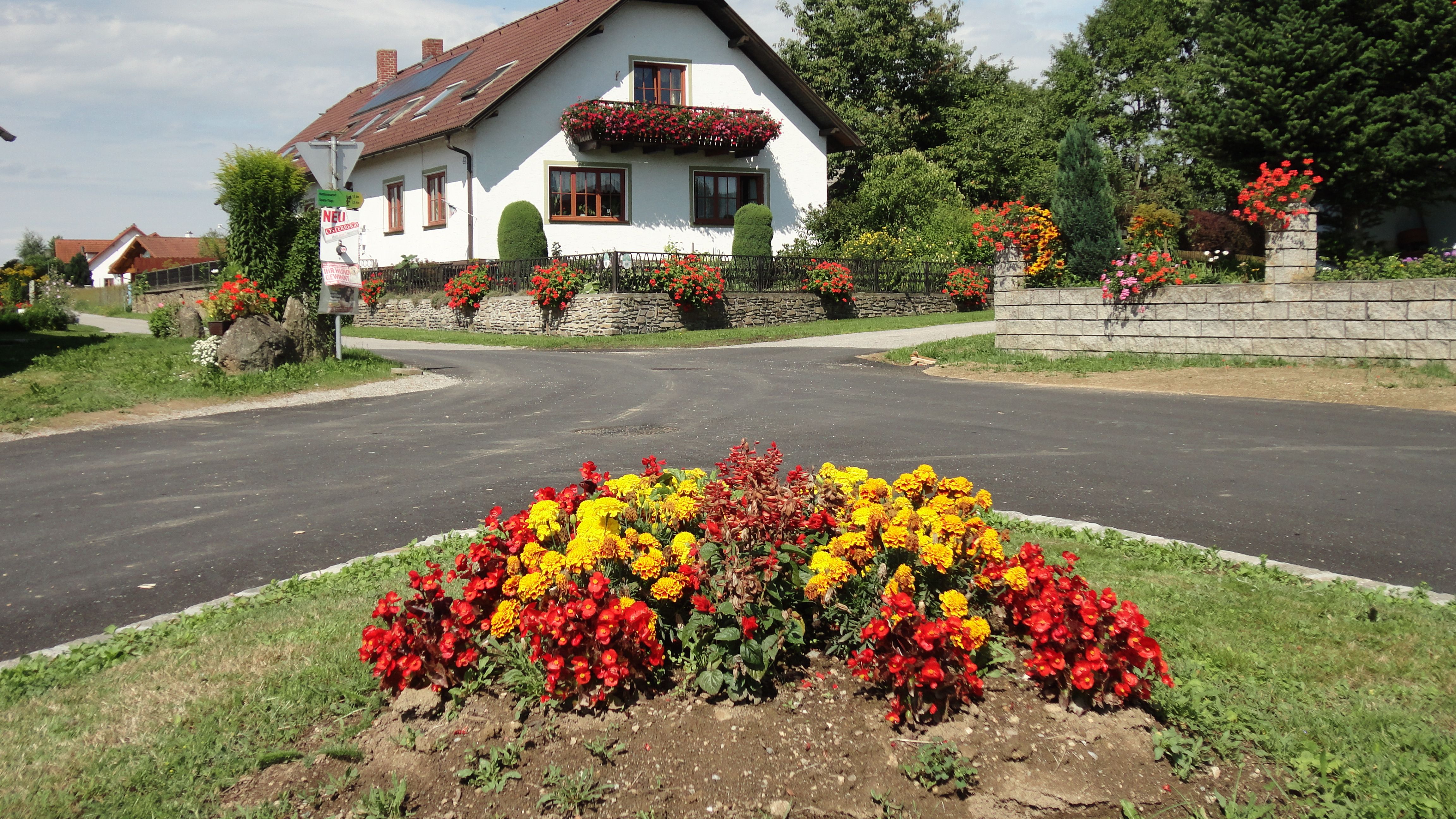 Ein weißes Haus mit roten Dachziegeln und Blumenschmuck, umgeben von bunten Blumenbeeten und grüner Landschaft.