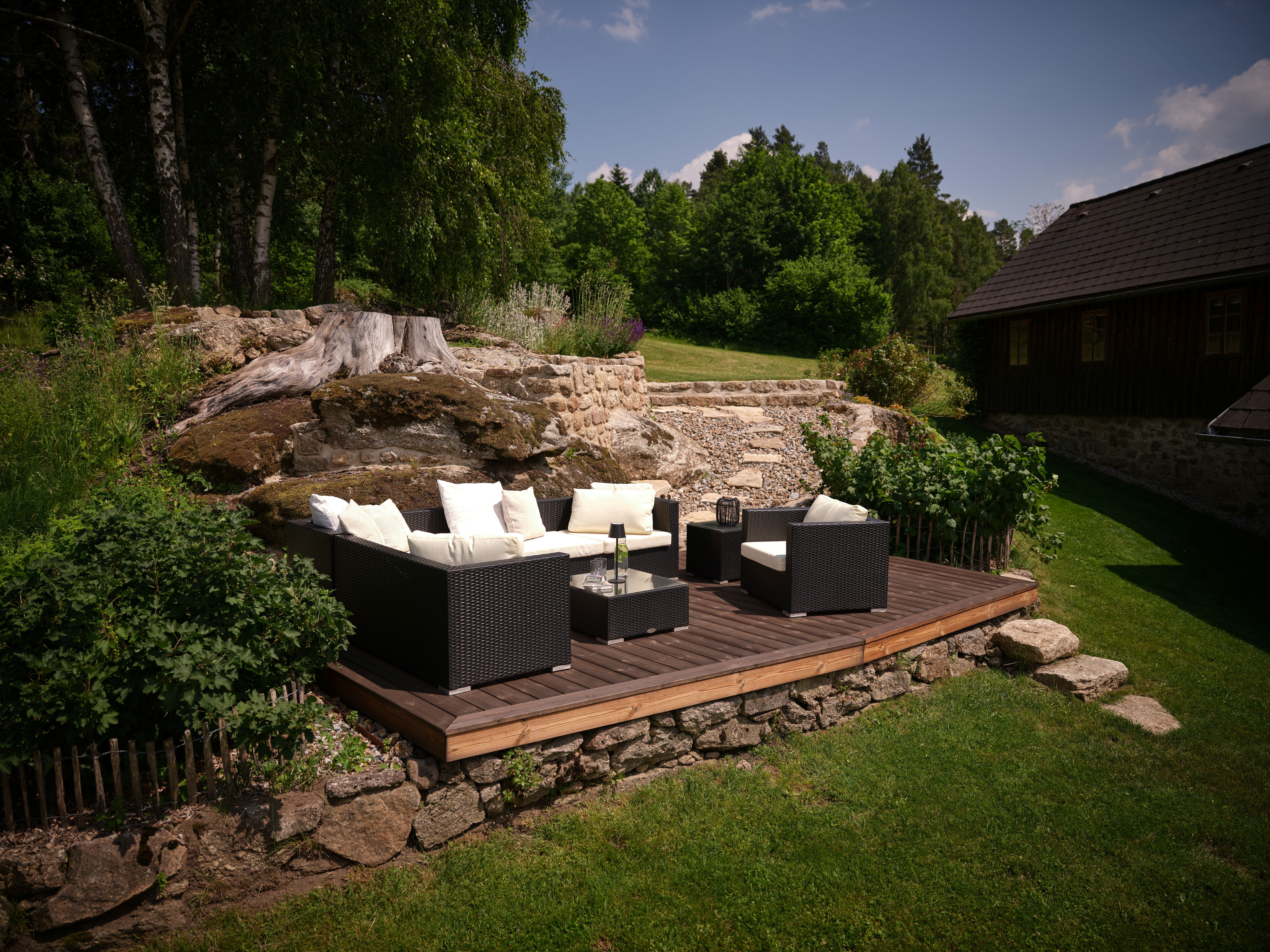 Garten mit Terrasse und Sitzmöbeln vor einem bewaldeten Hintergrund.
