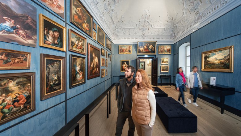 Besucher betrachten Gem&auml;lde in einem kunstvoll dekorierten Raum mit blauen W&auml;nden und verziertem Deckenstuck.