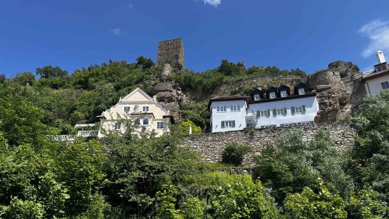 Blick auf Dürnstein mit historischen Gebäuden und Ruinen auf einem Hügel, umgeben von grüner Vegetation und blauem Himmel.