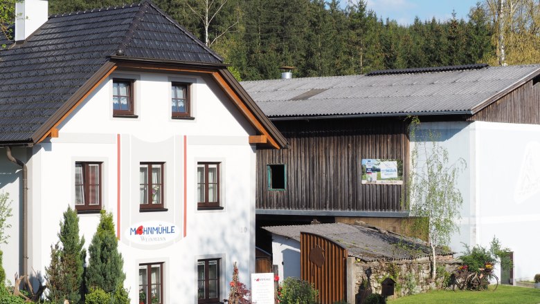 Geb&auml;ude der Mohnm&uuml;hle Weinmann mit angrenzendem Holzschuppen, umgeben von B&auml;umen.