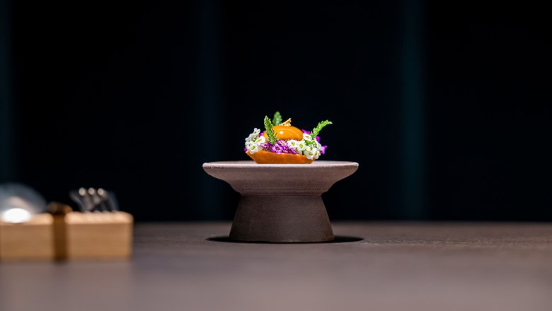 Ein kunstvoll angerichtetes Gericht auf einem kleinen Podest, umgeben von Blumen und Kr&auml;utern, vor dunklem Hintergrund.