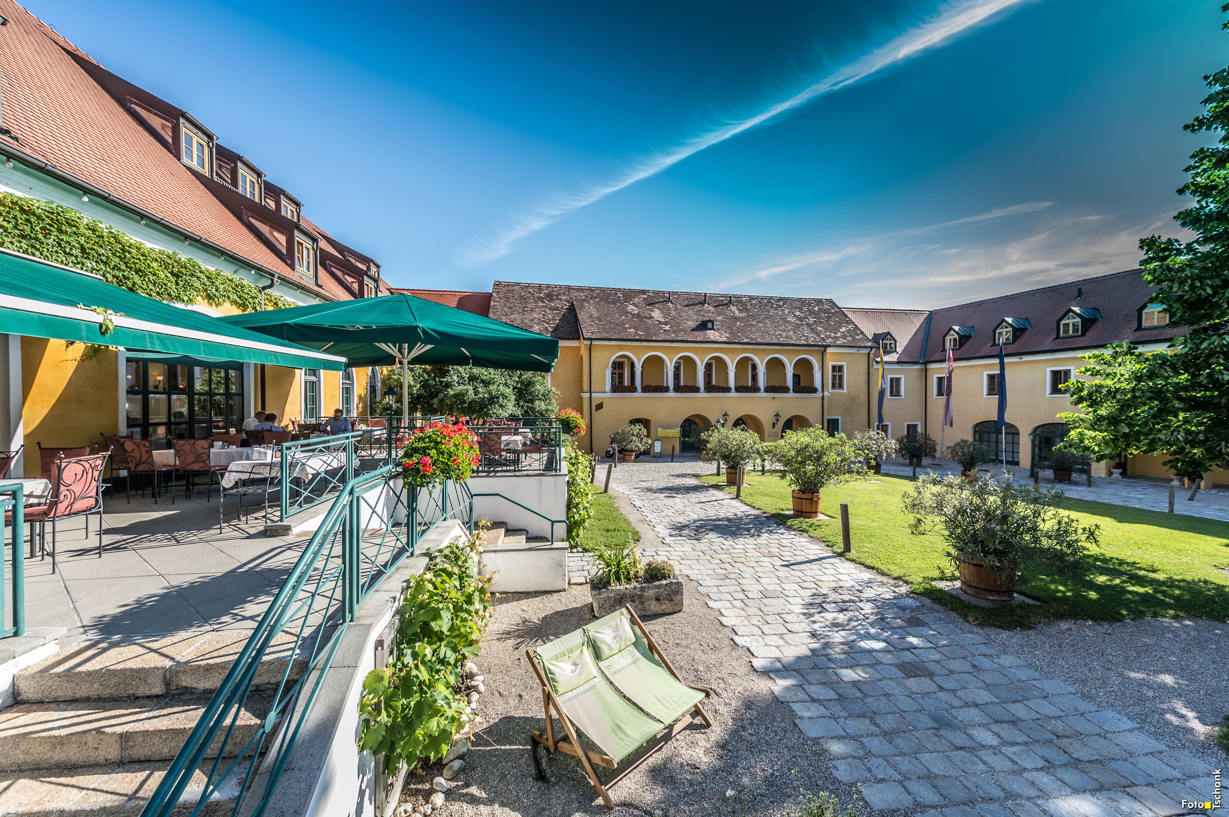 Ein sonniger Innenhof mit gelben Gebäuden, Terrassenbereich und grünen Sonnenschirmen.