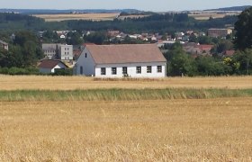 Ein weißes Haus mit rotem Dach steht in einem Feld, im Hintergrund eine Stadt und Wälder.