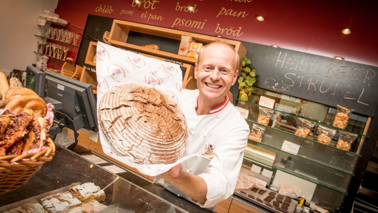 Ein Bäcker in einer Bäckerei hält ein großes, rundes Brot lächelnd in die Kamera. Im Hintergrund sind Regale mit Backwaren und eine Tafel mit der Aufschrift 'Heidelbeer Strudel'.