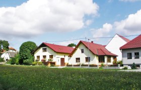 Ein Biohof mit mehreren gelben Gebäuden und roten Dächern, umgeben von grünen Wiesen und Bäumen unter einem blauen Himmel mit Wolken.
