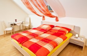 Helles Schlafzimmer mit rotem Bettbezug und dekorativen Vorh&auml;ngen.