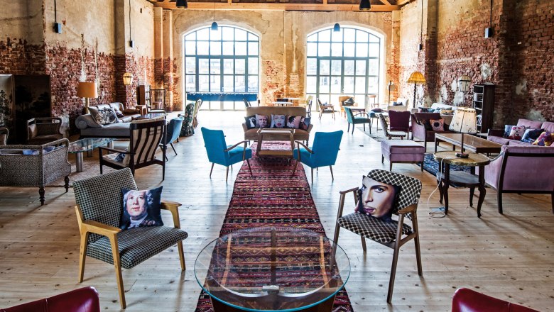 Innenansicht einer stilvoll eingerichteten Scheune mit Holzboden, gemauerten Wänden und großen Fenstern. Verschiedene Sitzmöbel und bunte Kissen sind zu sehen.