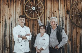 Three people in front of a wooden wall with wagon wheels, one person in cooking clothes.