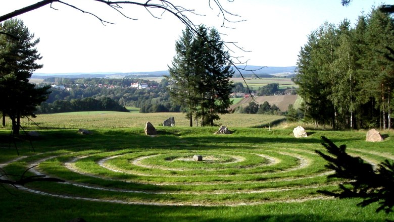 Ein spiralförmiges Labyrinth aus Gras und Steinen auf einer Wiese mit Bäumen und einem Dorf im Hintergrund.