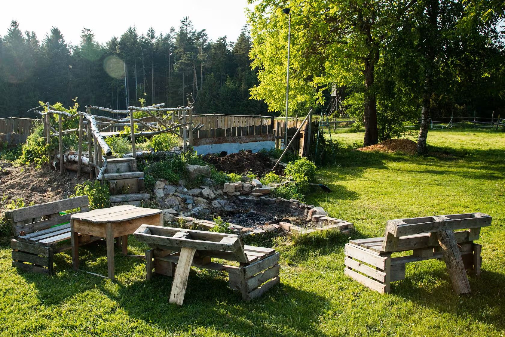 Rustikaler Garten mit Holzsitzgruppen, Feuerstelle, Bäumen und Wiese im Sonnenlicht.