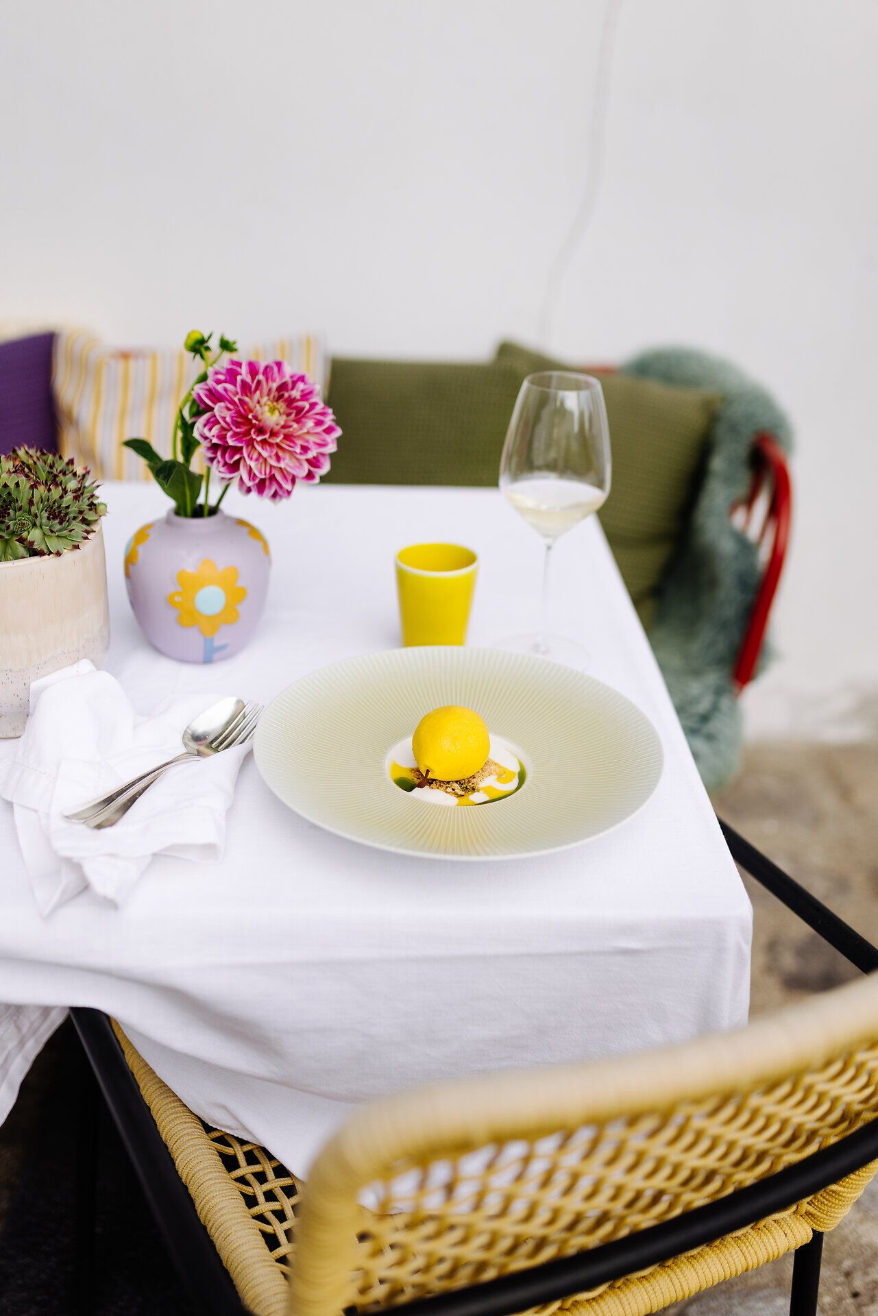Ein einladender Tisch im Freien, geschmückt mit bunten Kissen und einer blühenden Pflanze, lädt zum Verweilen ein. Auf dem Teller glänzt eine exquisite Nachspeise, die den Gaumen verwöhnt und die Sinne erfreut. Ein Glas Wein steht bereit, um den Moment in dieser charmanten Umgebung zu genießen.