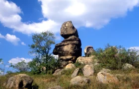 Ein markanter Felsen, umgeben von Büschen und Steinen, unter einem blauen Himmel mit weißen Wolken.