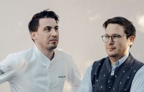 Zwei Männer stehen nebeneinander, einer trägt eine Kochjacke, der andere eine Trachtenweste.