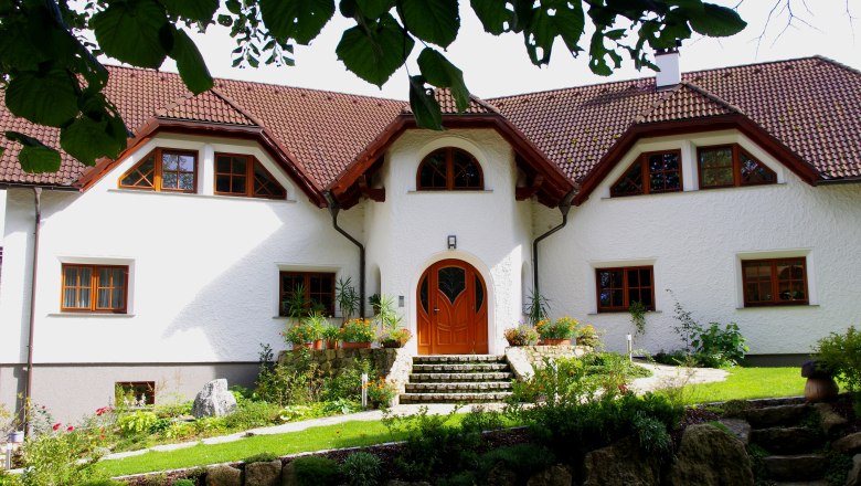 Ein großes, weißes Haus mit rotem Ziegeldach und gepflegtem Garten, umgeben von Bäumen.