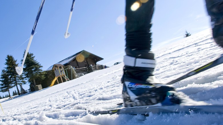 Nahaufnahme eines Langl&auml;ufers auf einer schneebedeckten Piste mit einer Holzh&uuml;tte im Hintergrund.