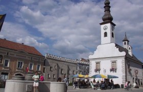 Ein Stadtplatz mit einem weißen Gebäude mit Turm und Uhr, Menschen sitzen unter Sonnenschirmen, ein Brunnen im Vordergrund.